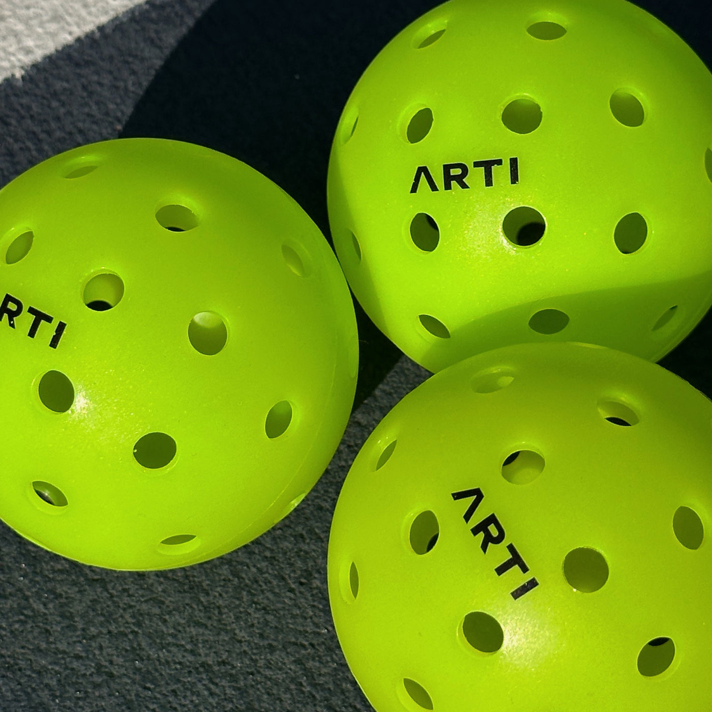 Three green ARTI outdoor pickleballs on a court in sunlight