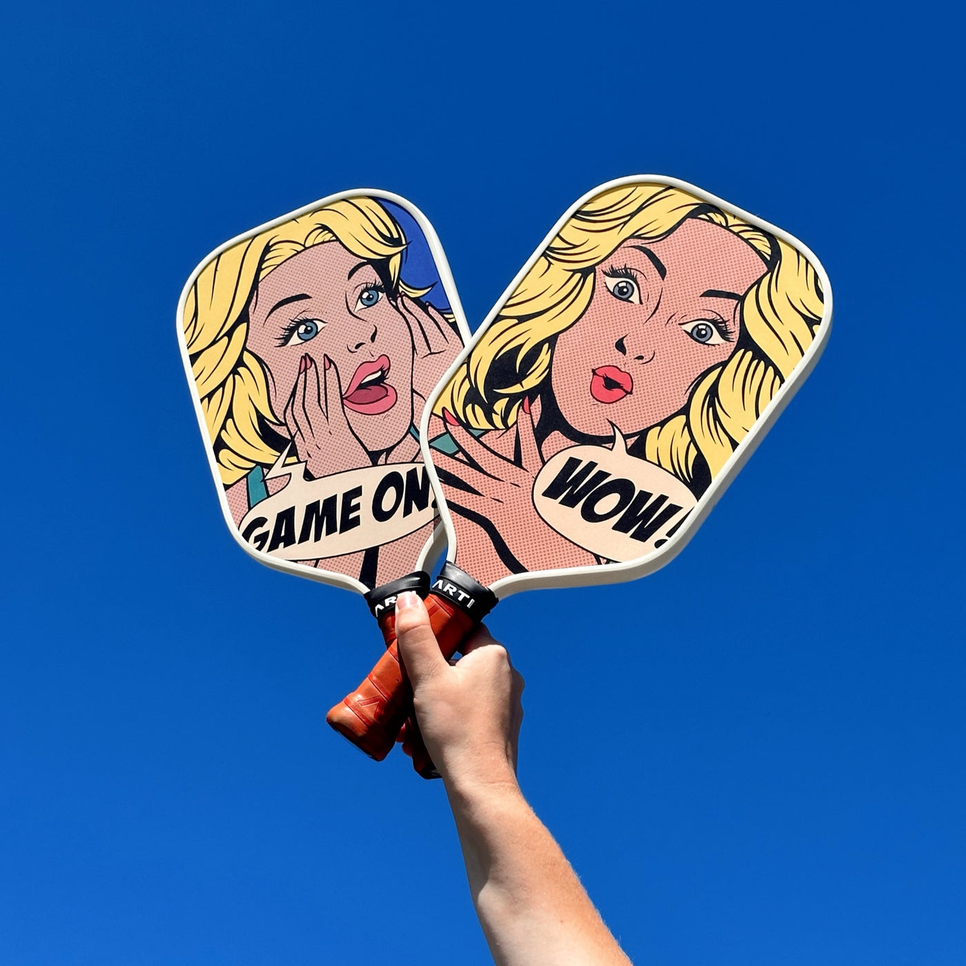 ARTI Game On K&K pop-art pickleball paddle held up against a blue sky